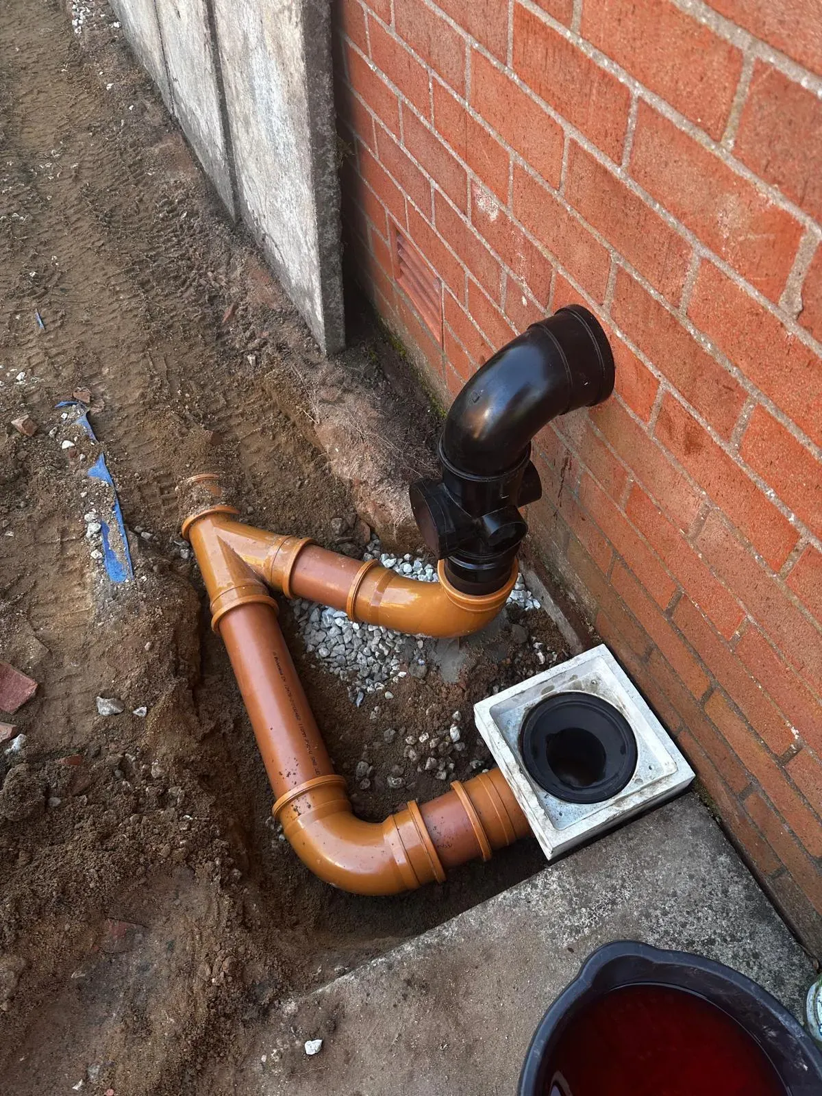 New orange and black plastic drainage pipes installed in an excavated trench next to a brick house wall, connecting to an external gully, demonstrating drain installation services.