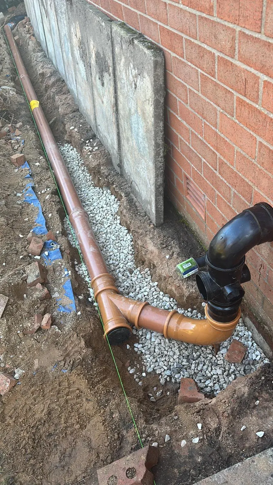 An image showing newly installed orange and black drainage pipes laid in a gravel bed within an excavated trench next to a brick building, indicating drain installation in progress.